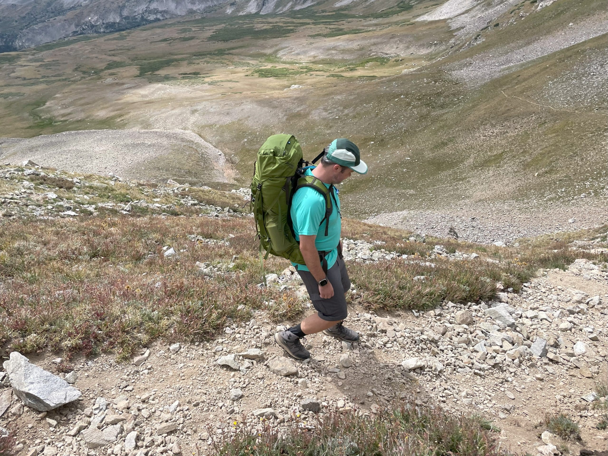 A man hiking down a steep, rocky hill while wearing the Osprey Aether 55 backpack in garlic mustard green.