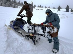 A man wearing the Patagonia Insulated Powder Town jacket and pants while pulling snow surfers off a snowmobile.